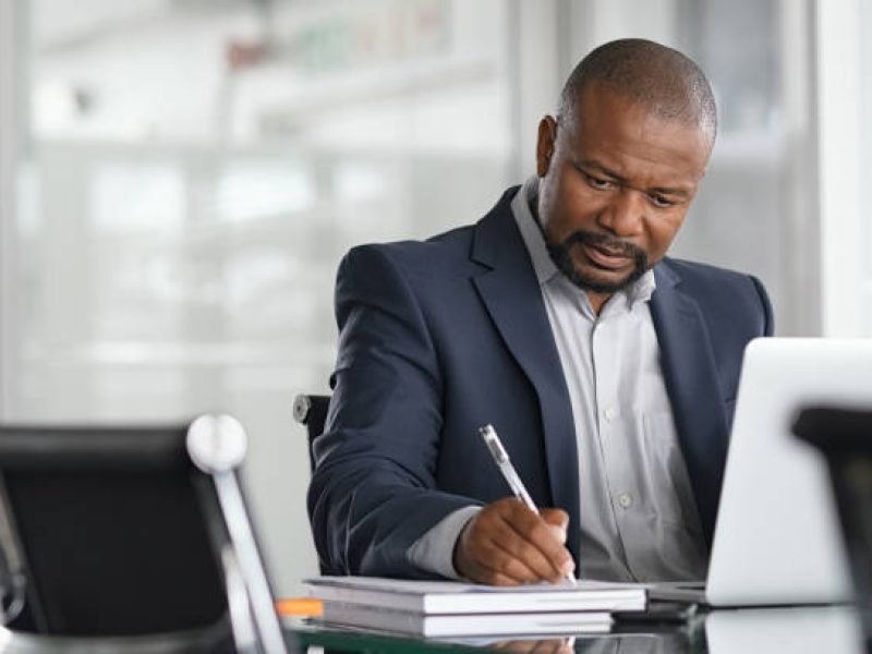 African serious businessman writing notes and using laptop. Mature business man writing his strategy on notebook while using laptop in modern office. Focused black entrepreneur sitting at desk in modern office while working.